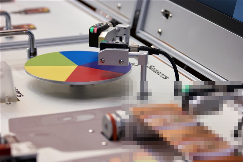 Industrial machine testing colour output with spectrophotometer sensors, measuring printed sheet accuracy against reference values.