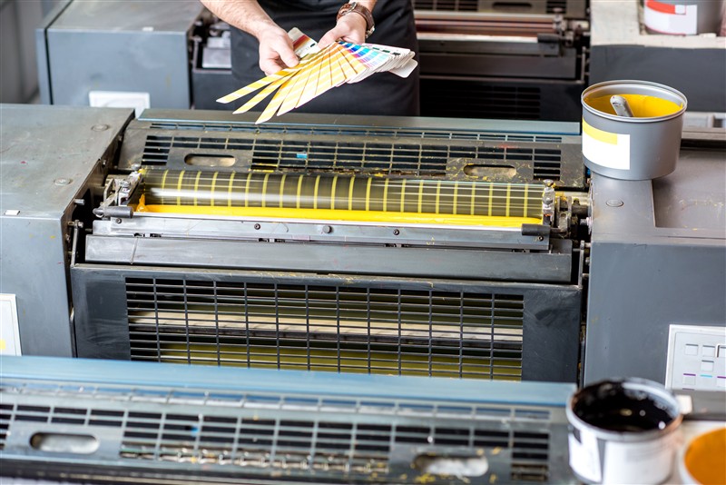 A technician monitors a sheet-fed offset press, checking registration and ink density as printed sheets stack in the delivery unit.