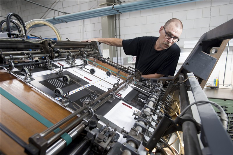 A worker monitors a colour sheet-fed press in a clean industrial workshop, reviewing the output at the delivery stack.